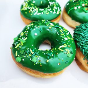 A St. Patrick’s Day-themed donut with green icing and shamrock sprinkles, decorated with a festive mix of green, gold, and white toppings.